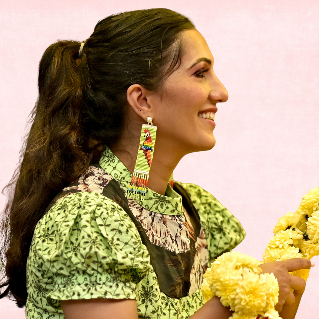 Woman wearing colorful earrings and a green patterned top against a pink background